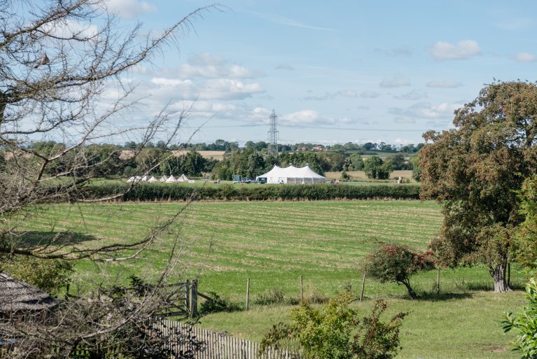Three pole wedding marquee set up in the distance with bell tents in the field for festival vibes at the wedding.