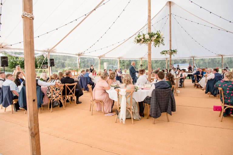 Wedding guests inside the large sailcloth tent with flower hoops around the poles and round tables and chairs. Guests toasting the speeches in the structure.