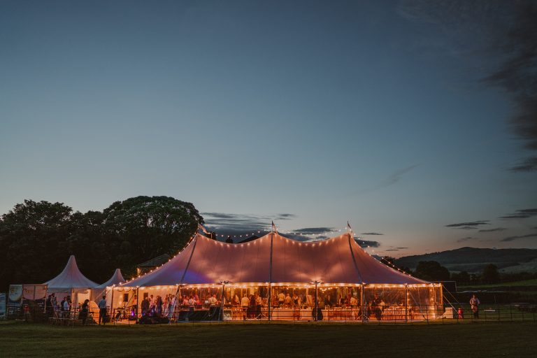Wedding marquee lit up for a evening party with festoon lights over the top of the tent and up lighters around the marquee. Structure in the grounds of Broughton Hall Yorkshire.