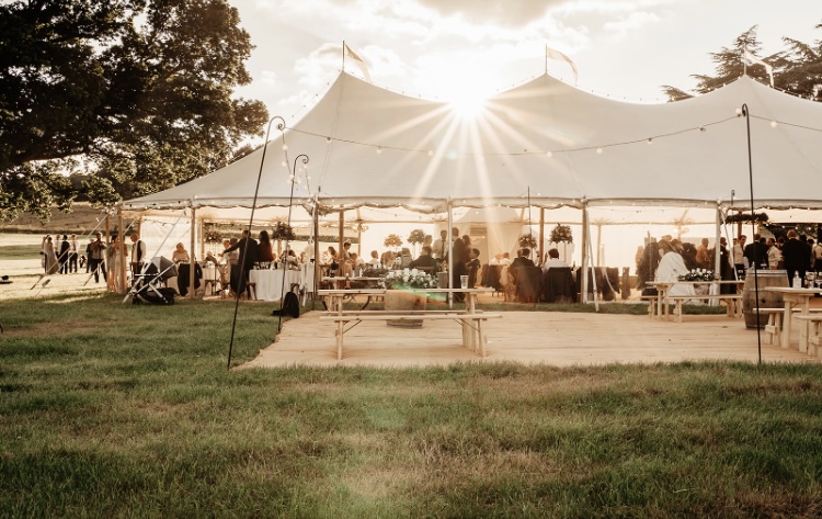Large Sperry style tent at sunset with wedding guests around and flags on top of the structure.
