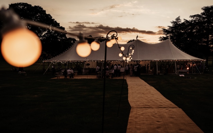 Evening in the sailcloth tent, festoon lights along the walkway. Large Aurora Sailcloth tent with 4 poles for a wedding party in the grounds of a stately home in Yorkshire.