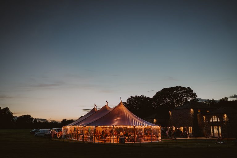 Sailcloth marquee with three poles in the tent. Wedding party of 150 guests in the marquee in Yorkshire. Lit up in the evening with festoon lights over the tent and the marquee structure is in the grounds of an estate in Yorkshire.
