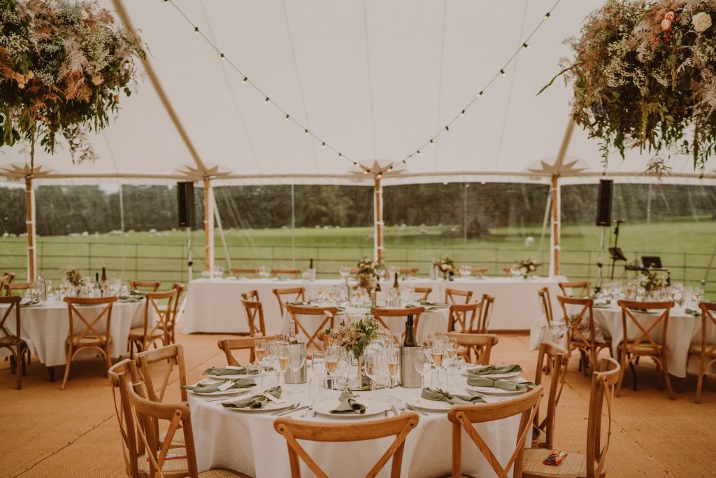 Interior of the sailcloth tent laid up with round tables and chairs for the wedding. Flower hoops around the poles and matted flooring.