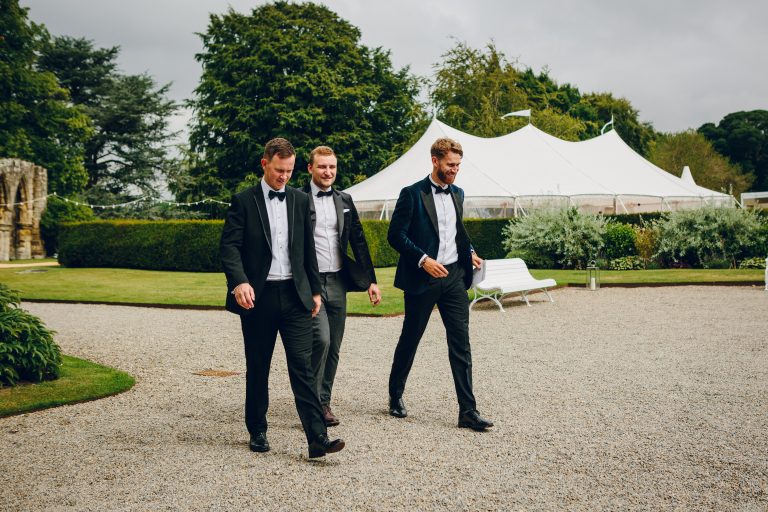 pole tent with three poles and flags set up in the grounds of a stately home. Best men walking in front of the pole tent for the wedding
