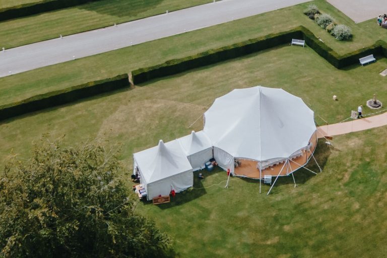 pole tent set up in the grounds of a stately home. pole tent to be used for a wedding. festoon lights along the matted walkway. round tables inside the marquee. catering marquee connected to the main marquee with a link. clear sides around the pole tent