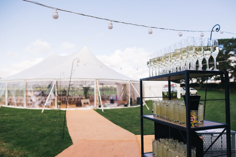 Matted walkway and festoon lights up the marquee. drinks trolley with champagne glasses on it at the entrance of the marquee
