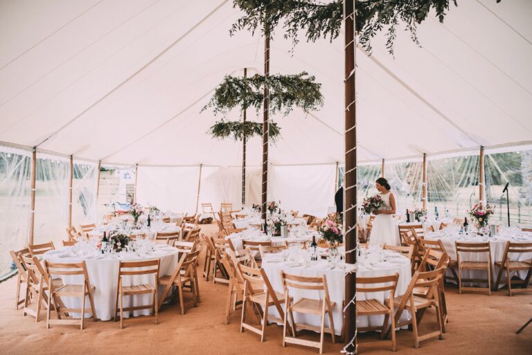 Interior of the sailcloth tent laid up with round tables and chairs for the wedding. Flower hoops around the poles and matted flooring.