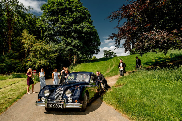 Wedding car being pushed down a hill outside the church with the best men pushing the car and the bridesmaids watching