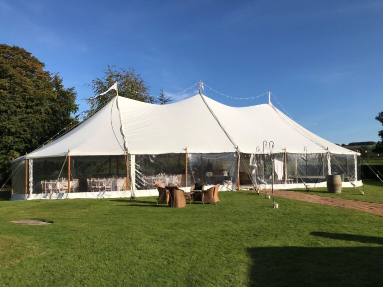 pole tent exterior with rattan chairs and table outside the marquee and matted walkway to the entrance. festoon lights over the top of the pole tent blue skies