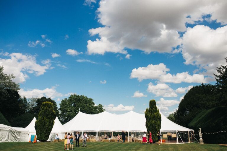 blue skies and pole tent with four peaks. white canvas and clear sides. grass surrounding the marquee with a few people